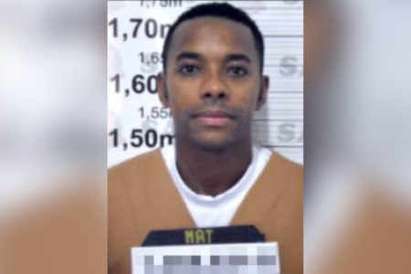 Imagem colorida mostra o ex-jogador Robinho, um homem negro de cabelos curtos, vestindo uniforme de presidiário, posando para foto na cadeia - Metrópoles