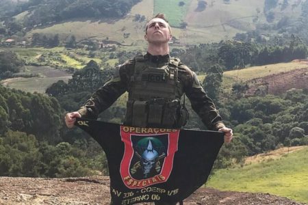 Foto colorida de policial com farda verde escura, em montanha em dia ensolarado, segurando abaixo da cintura uma bandeira do grupo de operações especiais da PM de São Paulo - Metróles