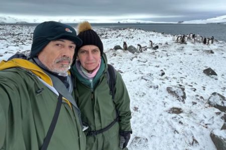 Foto mostra um homem e uma mulher em uma paisagem nevada com centenas de pinguins atrás deles- Metrópoles gripe aviária