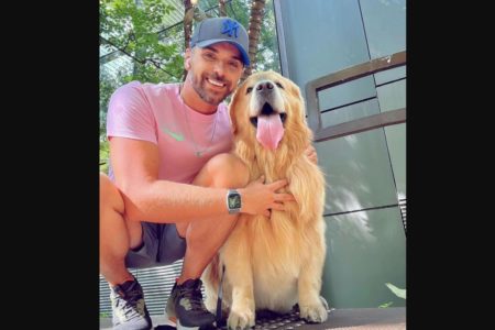 Homem branco com bermuda, camiseta rosa e boné azul está agachado ao lado de cão da raça Golden Retriever