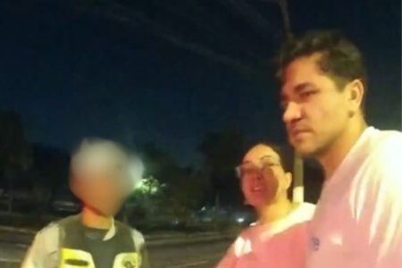 Imagem colorida de homem branco, ao lado de mulher branca de óculos, ambos com camiseta branda ao lado de PM na rua - Metrópoles