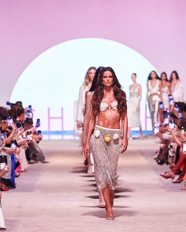 Izabel Goulart na passarela de desfile com outras modelos atrás. A modelo usa um top claro tridimensional e uma saia com transparência - Metrópoles