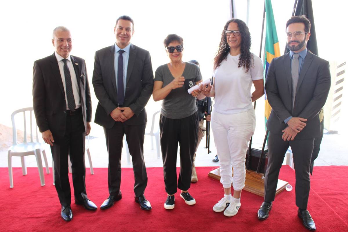 Foto oclroida de cinco pessoas, sendo quatro autoridades e uma quinta mulher trans presa