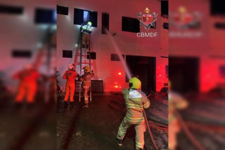 Foto colorida tirada à noite de casa iluminada por luz externa vermelha e homem com uniforme de bombeiro operando uma mangueira d'água para apagar chamas