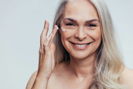 Foto colorida de uma mulher de cabelo grisalho sorrindo enquanto passa creme no rosto com os dedos - Metrópoles