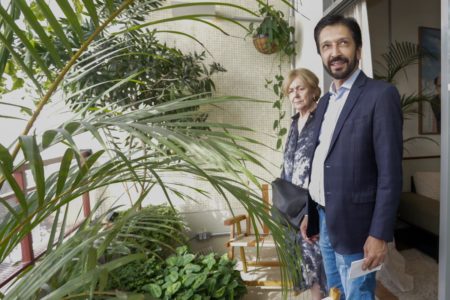 Imagem colorida mostra Ricardo Nunes, homem branco, de cabelo e barba pretos, vestindo terno azul, camisa branca e calça jeans, em uma sacada de prédio decorada com plantas - Metrópoes