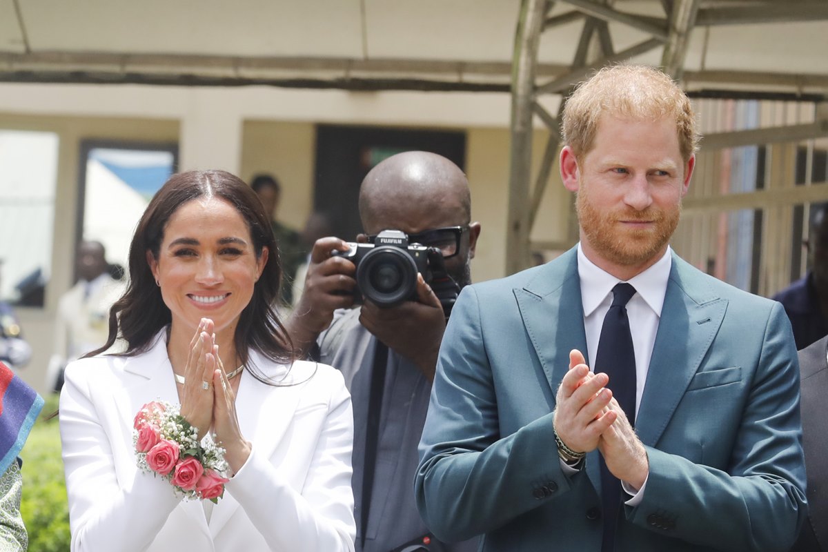 Foto colorida de mulher com roupa branca ao lado de homem com ruivo com blazer e gravata azul. No meio deles, há um homem com uma máquina fotográfica em frente ao rosto - Metrópoles