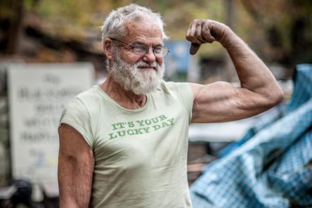 Idoso barbudo, de aparência robusta, com mãos sujas, vestindo camiseta humorística e exibindo o músculo bíceps flexionado. Isoso musculoso, forte, com testosterona