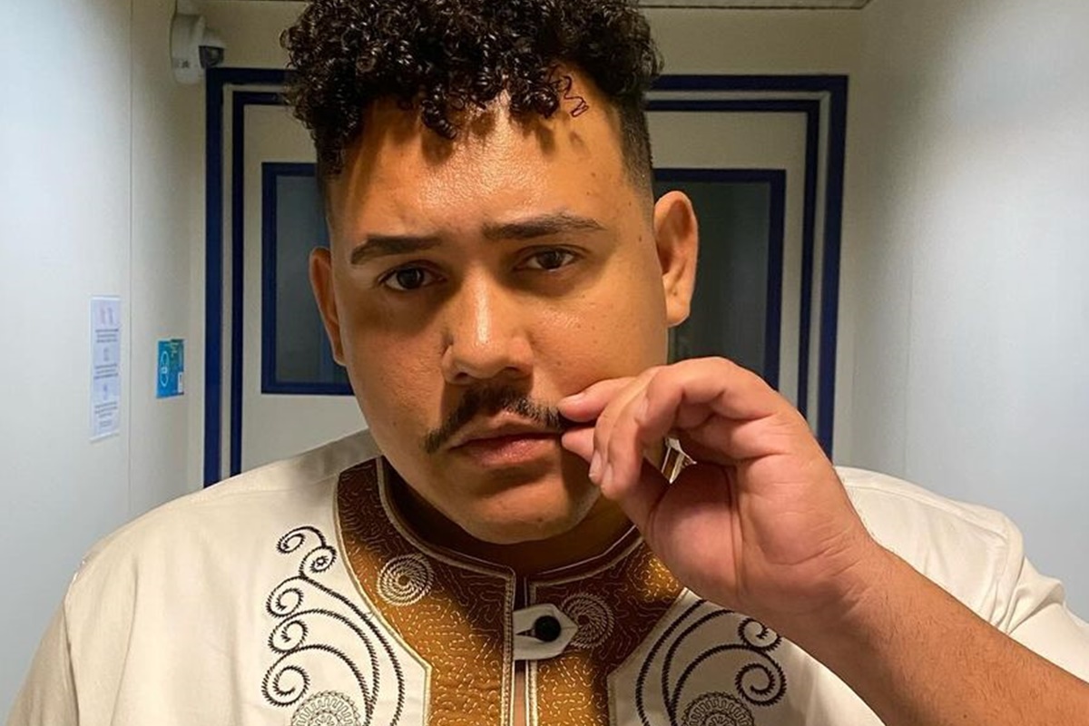 O ex-BBB Lucas Henrique, o Buda, posa com umablusa branca e marrom, segurando o bigode - Metrópoles