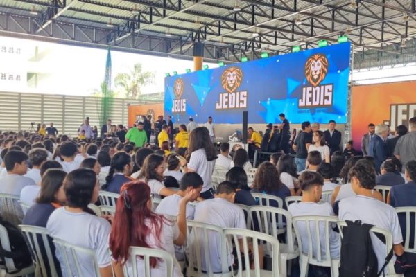 Foto colorida de estudantes de camiseta branca sentados em evento esportivo