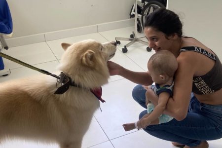 Foto colorida de mulher com bebê no colo fazendo carinho em um cachorro peludo