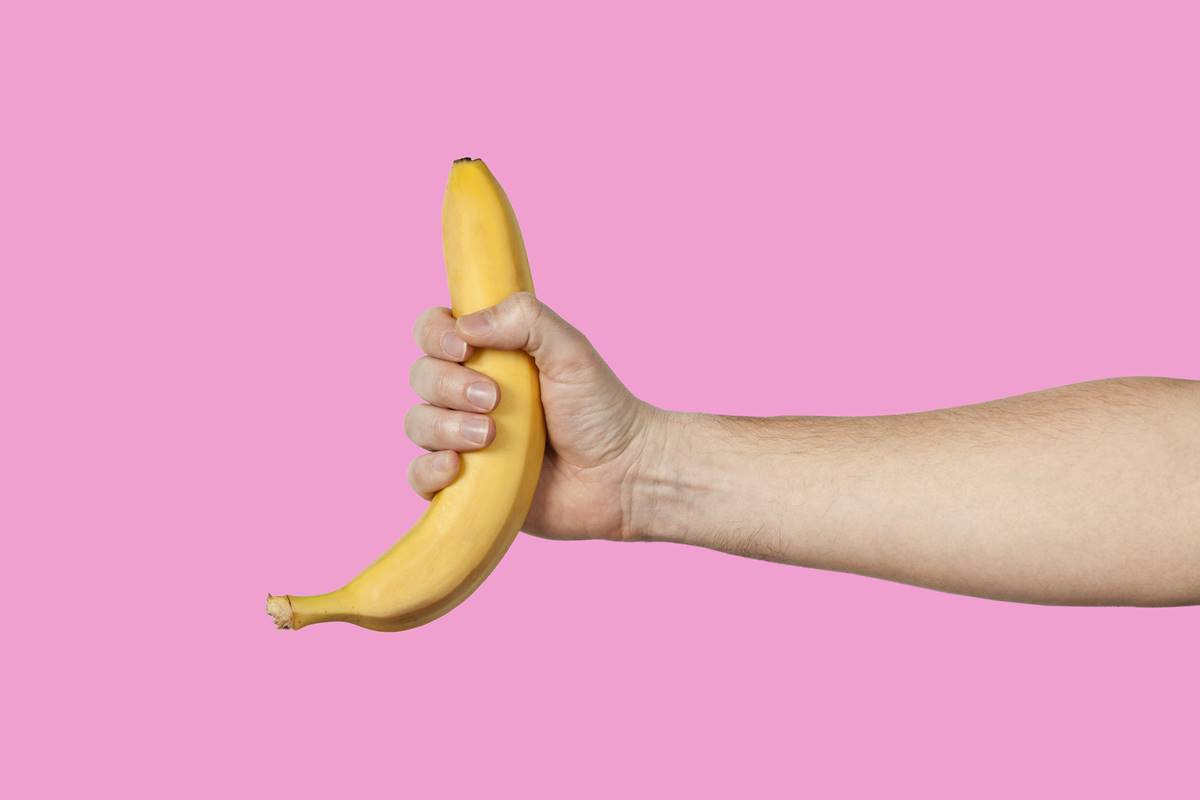 foto colorida de uma mão masculina segurando uma banana com casca em um fundo cor-de-rosa - Metrópoles
