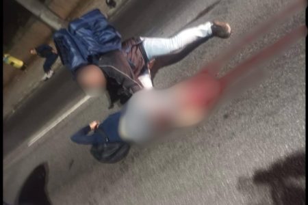 Imagem colorida, com desfoque da cintura para baixo de mulher caída em asfalto sendo amparada por motoqueiro com mochila de entrega azul - Metrópoles