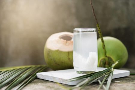 Foto colorida - Copo com água de coco e coco verde atrás