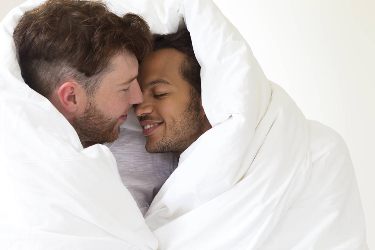 Foto colorida de um casal com os rostos próximos e sorrindo embaixo de um adredom branco - Metrópoles