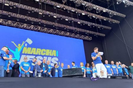 imagem colorida mostra grupo de pessoas em cima de palco durante evento da marcha para jesus. atrás deles, um telão exibe o nome do evento.