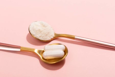 Foto colorida - Duas colheres douradas em um fundo rosa, uma com colágeno em pó e outra com colágeno em capsula