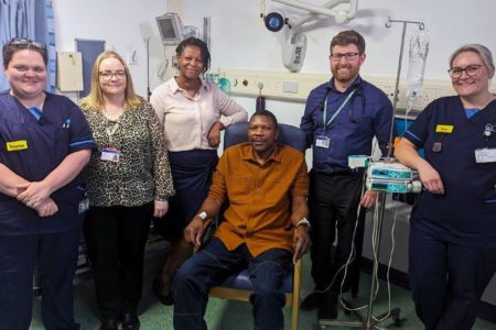 Foto mostra Elliot Phebve de camiseta laranja sentado ao lado da equipe médica e de sua esposa. Ele é o primeiro vacinado do mundo contra o câncer de intestino - metrópoles
