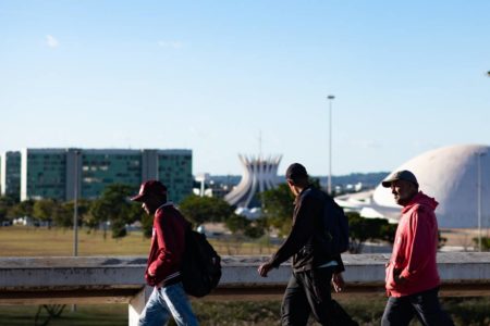 Foto colorida de três homens de roupa de frio caminhando em frente a pontos turísticos da Esplanada de Brasília