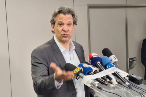 Imagem colorida mostra o ministro da Fazenda Fernando Haddad, um homem branco vestindo um terno cinza e uma camisa branca, diante de um púlpito com microfones de diversas emissoras Senado - Metrópoles