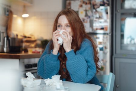 Imagem mostra mulher ruiva de blusa azul assoando o nariz. Vários papéis amassados estão na frente dela - Metrópoles