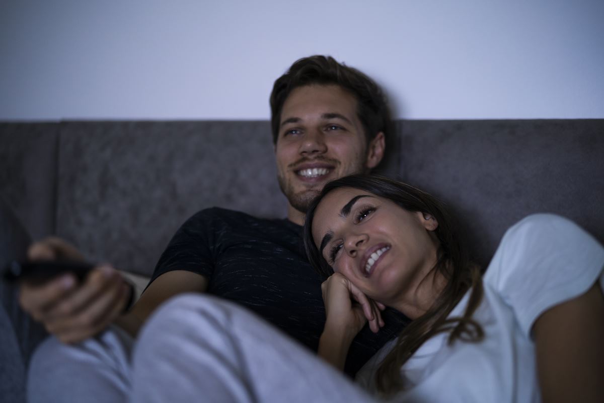 Foto colorida - Um homem e uma mulher sentados no sofá olhando para TV com o controle na mal