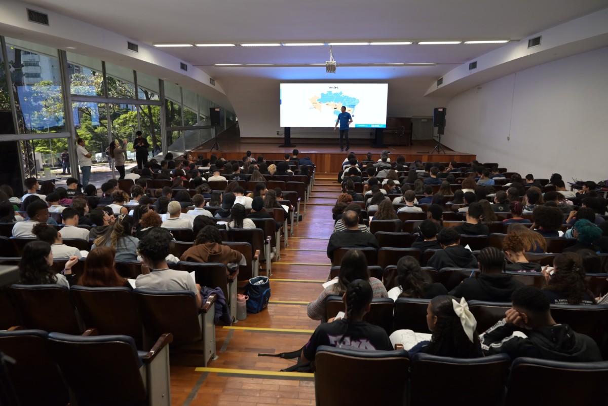 Fotografia colorida mostrando auditório cheio de alunos com professor em cima de palco dando aula na frente de telão mostrando mapa do Brasil-Metrópoles
