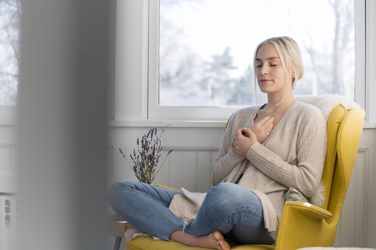 Mulher sentada no sofá de olhos fechados praticando exercícios de respiração ou meditação - Metróples