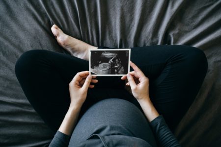 Foto de alto ângulo de uma mulher grávida segurando uma foto de ultrassom na frente de sua barriga, sentada na cama em casa. Maternidade - Metrópoles