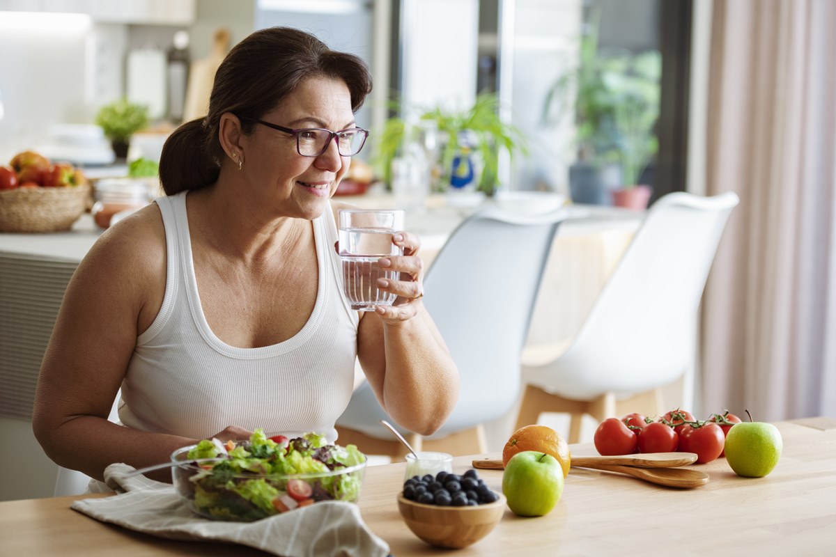 Foto colorida de mulher tomando água. Ela está próximo de um prato com salada - Metrópoles