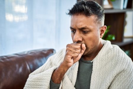 Foto de um homem tossindo enquanto se recuperava de uma doença no sofá de casa. Coqueluche - Metrópoles