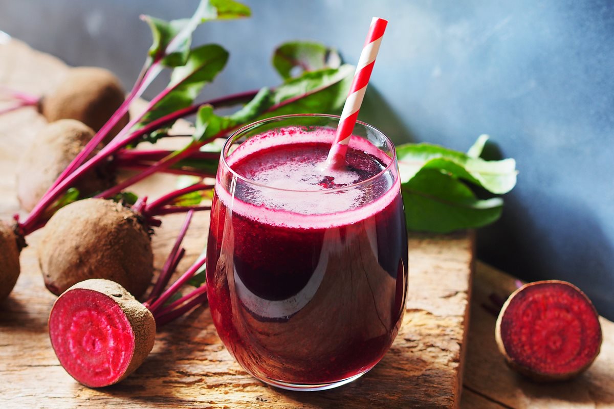 Copo de vidro com suco de beterraba e um canudo sob uma tábua de madeira. Metrópoles