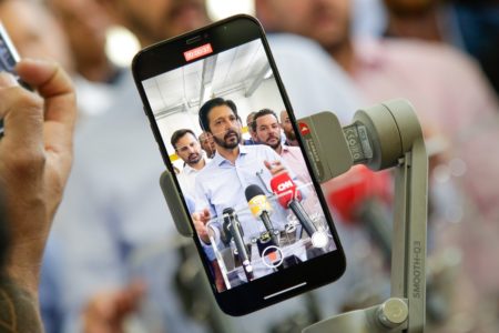 Imagem colorida mostra Ricardo Nunes, prefeito de SP, homem branco, de cabelo e barba pretos, vestindo camisa branca, na tela a de um celular - Metrópoles