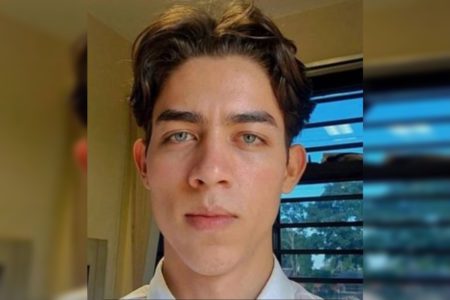 Imagem colorida de rosto de jovem branco, de cabelos castanhos lisos e curtos. Ele olha sério para a câmera e usa camisa branca