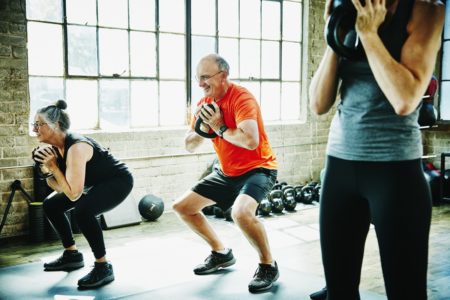 Atletas idosos e sorridentes fazendo agachamentos com kettlebell durante a aula de fitness - Idosos treinando, atividade física exercício idosos