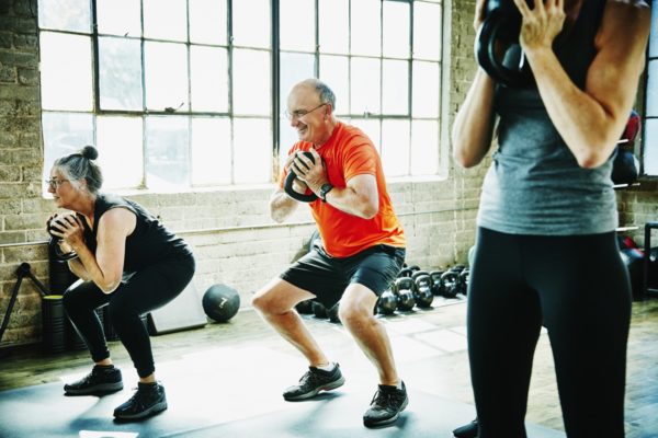 Atletas idosos e sorridentes fazendo agachamentos com kettlebell durante a aula de fitness - Idosos treinando, atividade física exercício idosos