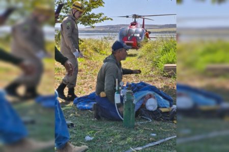 Imagem colorida de bombeiros militares socorrendo uma pessoa ferida, deitada sobre uma maca no chão, coberta com panos azuis