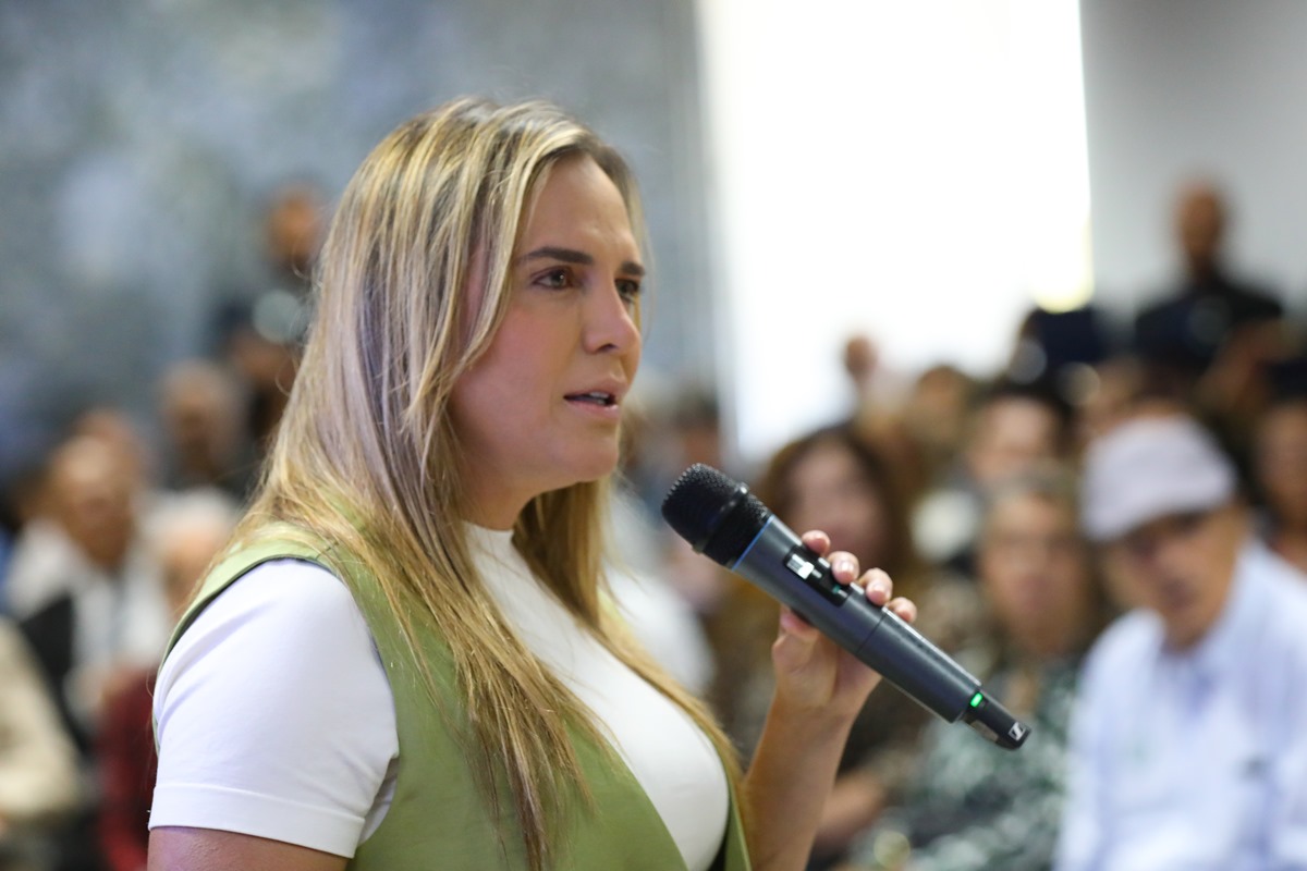 Foto colorida de mulher branca, loira, de cabelos lisos, blusa branca e colete verde falando ao microfone