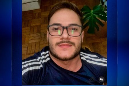 Imagem colorida de um homem branco, de óculos, barba, fazendo uma selfie. Ele veste camiseta azul com listras brancas nas mangas