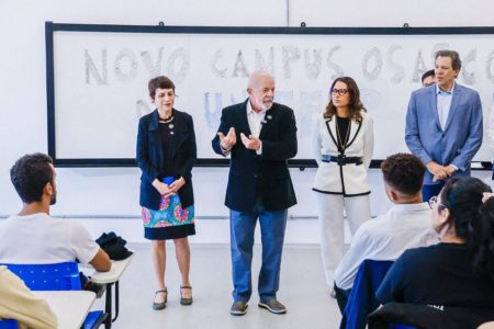 Homem branco de cabelos branco, blazer preto e calça jeans gesticula em sala de aula; ao fundo está o quadro, ao lado há outras pessoas e pé e, na frente dele, estudantes sentados em carteiras - Metrópoles