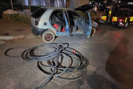 Foto colorida de carro abandonado em via de quadra residencial com vários cabos elétricos em volta