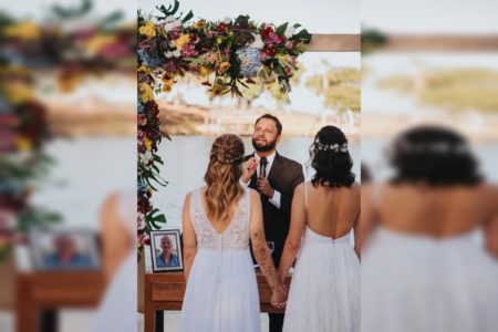 Imagem colorida de um casal de noivas, usando vestido branco, com o Pastor Henrique Vieira ao fundo. Ele usa um terno preto