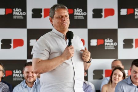 Imagem colorida mostra Tarcísio de Freitas, homem branco e grisalho, falando ao microfone. Ele veste uma camisa de mangas curtas clara e sorri enquanto fala ao microfone. Ao fundo, pessoas sentadas e um banner do governo de SP - Metrópoles