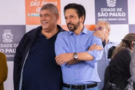 Imagem colorida mostra Milton Leite, homem negro, de cintura larga, vestido roupas escuras. abraçado ao prefeito Ricardo Nunes, homem branco, de cabelo e barba pretos, vestindo camisa azul - Metrópoles