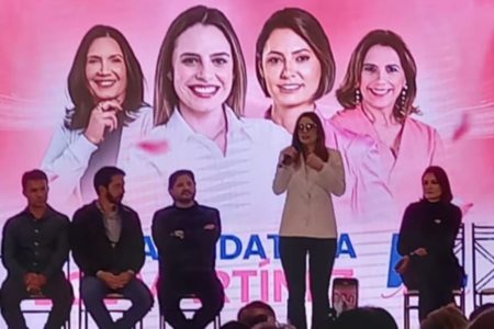 Foto colorida de Zoe Martínez em pé segurando microfone em palco com fundo cor de rosa e ao lado de políticos - Metrópoles