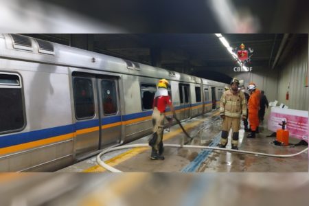 Imagem colorida de um trem de metrô passando por um incêndio, com três bombeiros vestidos de farda marrom atuando no combate