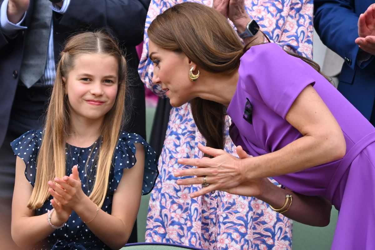 Foto colorida de menina loira batendo palmas e uma mulher com vestido roxo falando no ouvido da menina - Metrópoles