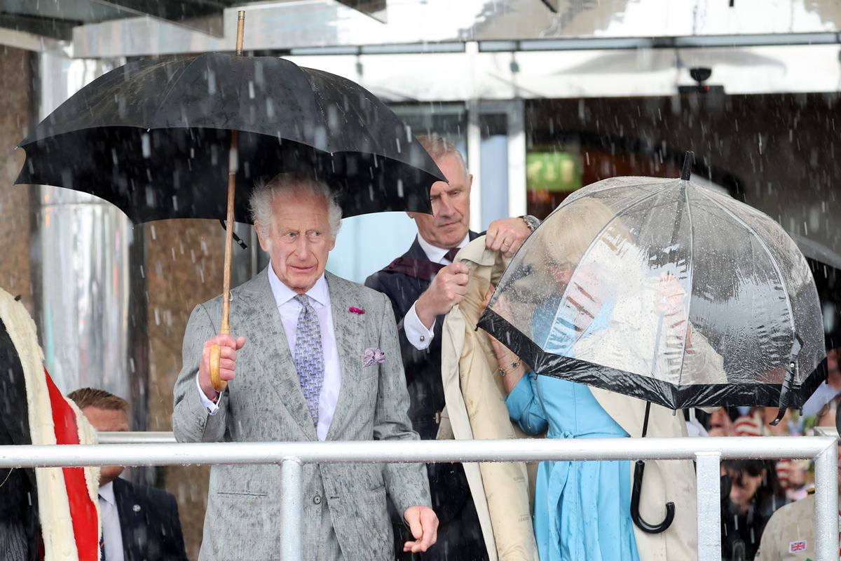 Foto de homem e mulher segurando guarda-chuva. Um homem ajuda a mulher a vestir uma capa - Metrópoles