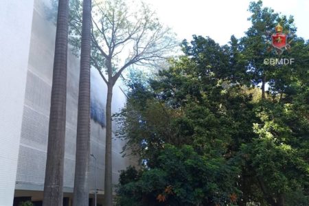 Foto colorida de prédio com fachada branca e fumaça saindo por cobogós. Árvores em frente ao edifício