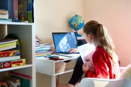 Menina sentada em frente a um computador em cima de uma mesas com caderno, caneta e folhas- Metrópoles